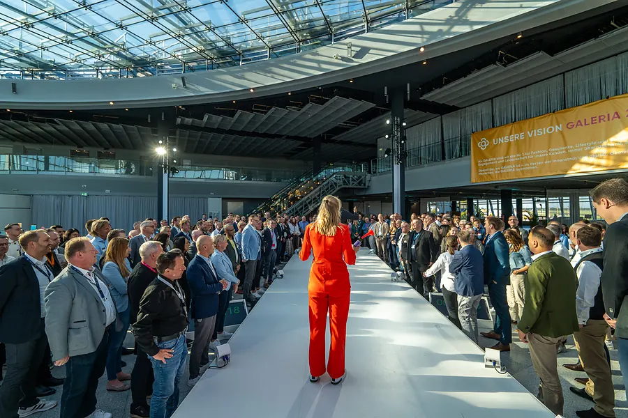 Desirée Ackermann im orangefarbenen Anzug auf dem Laufsteg vor vollem Saal